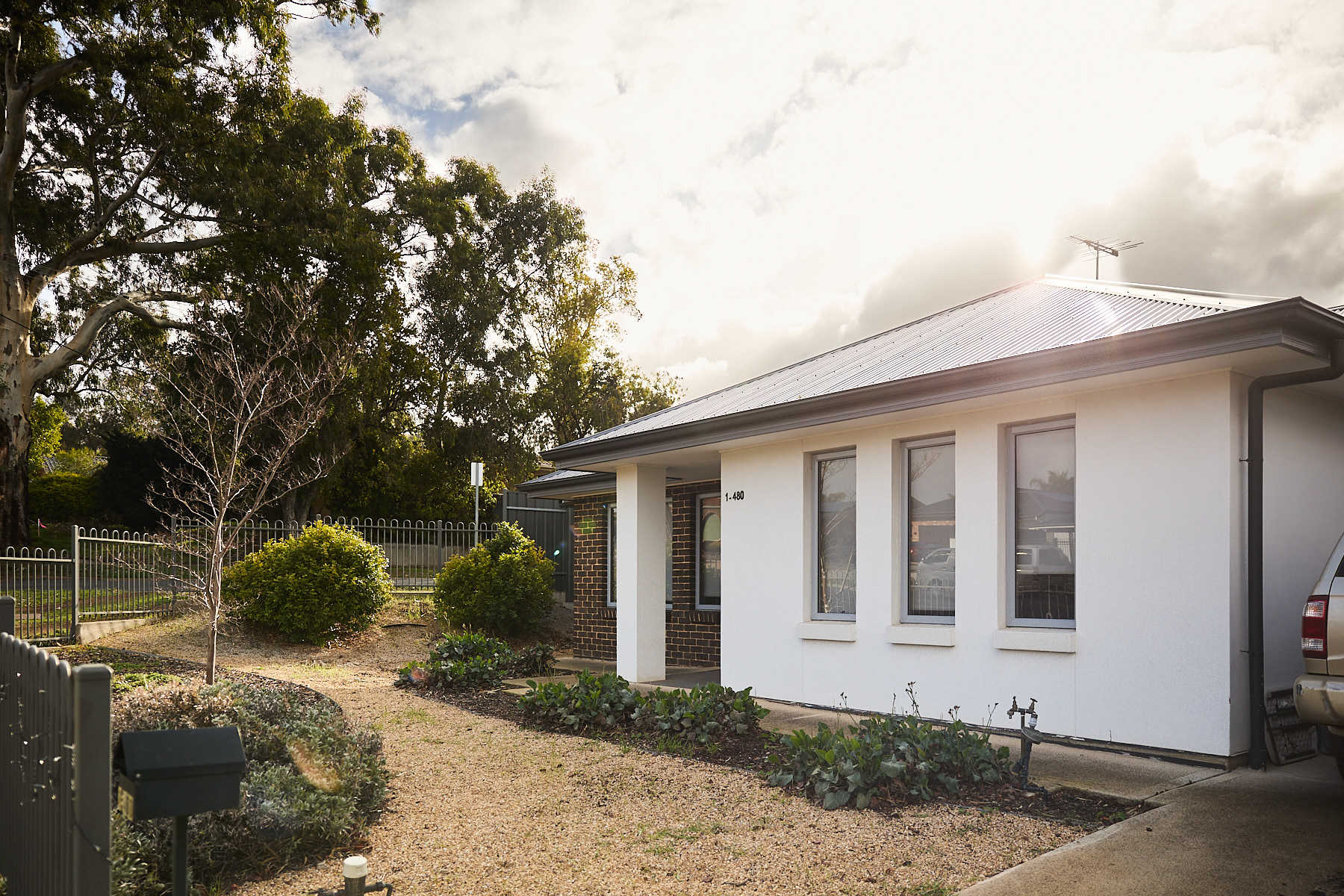 The image is of a small white house with a door on the left side and three windows to the right. The home has a gravel path and trees around it.