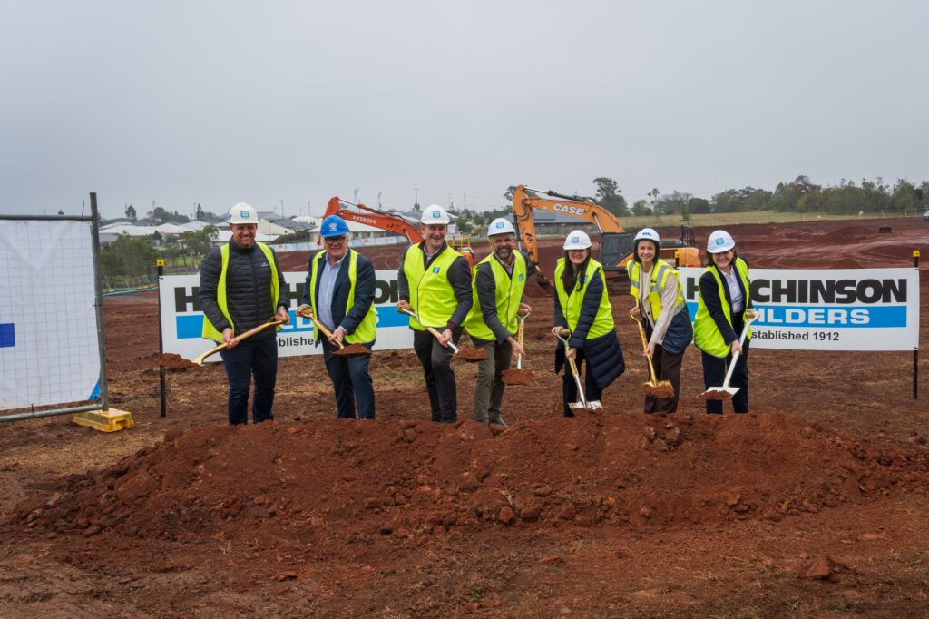A group of women and men wearing high vis and hard hats on a building site - they are using shovels to complete the official 'sod turn' at a new housing development.