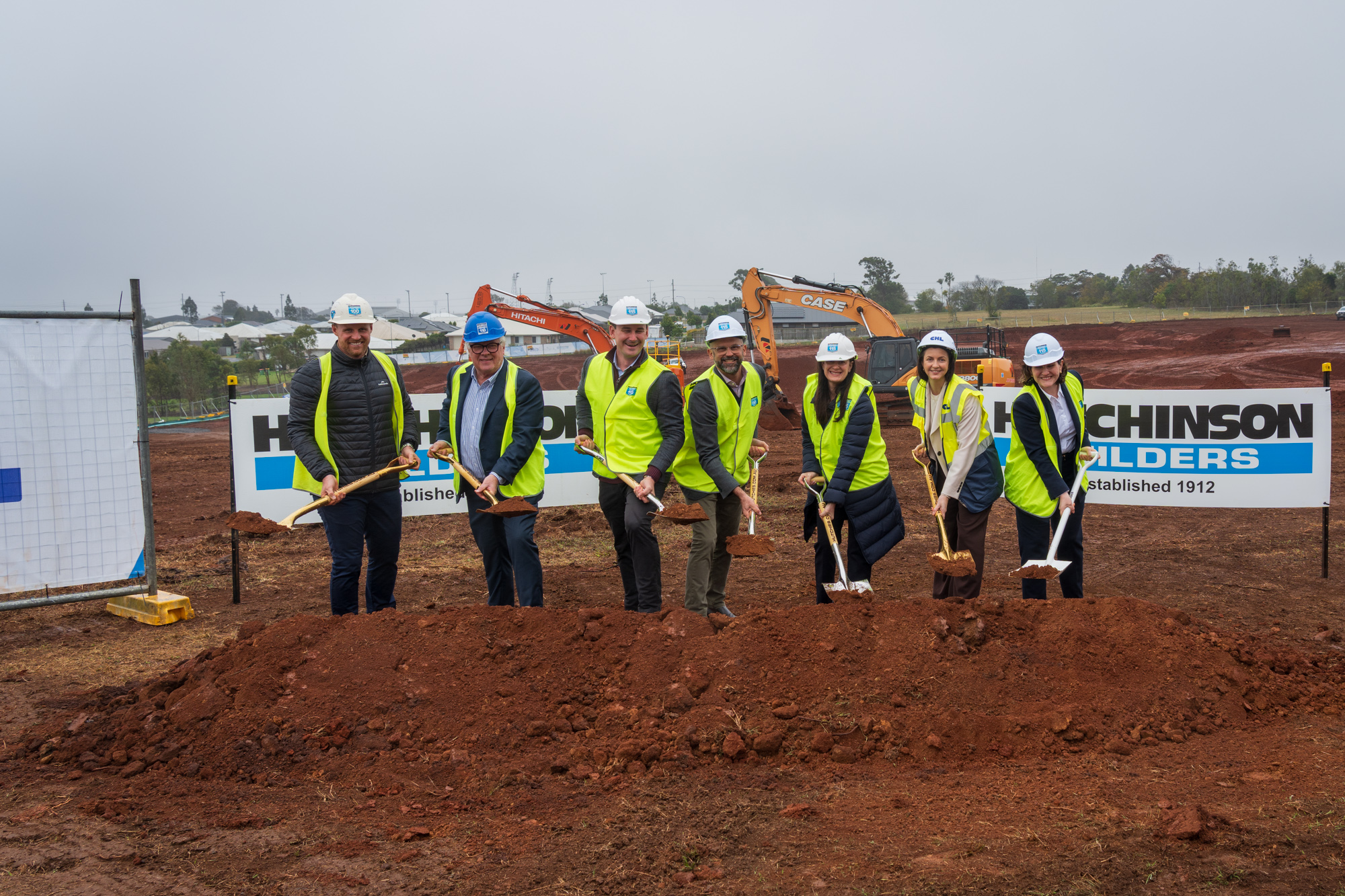 A group of women and men wearing high vis and hard hats on a building site - they are using shovels to complete the official 'sod turn' at a new housing development.