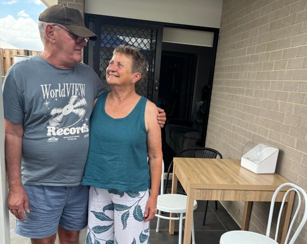 An older lady and man standing in front of their home, smiling at each other.