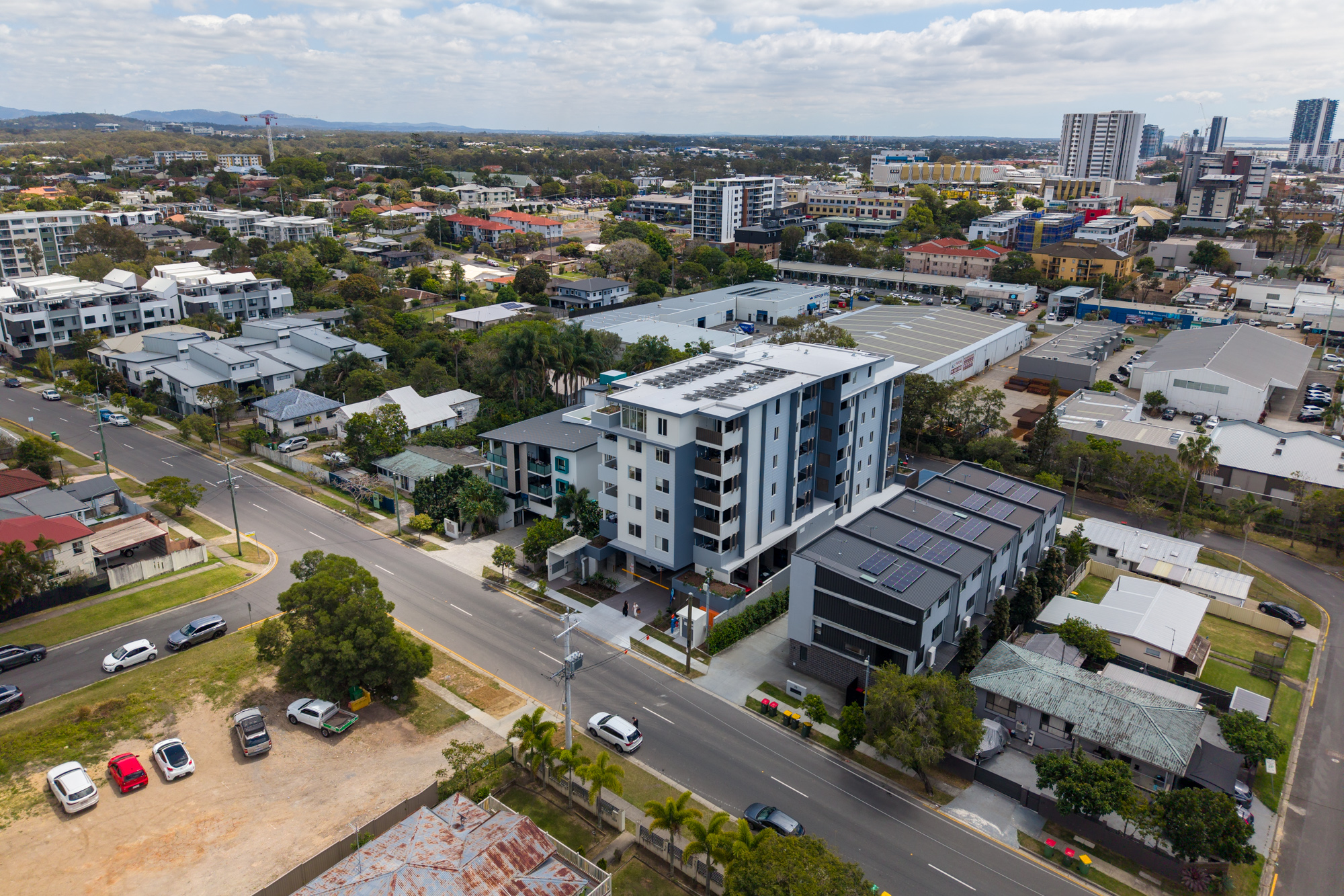 A multi-story apartment block aerial view of the apartments in the streetscape. Apartment block is white with windows and balconies, with landscaping out front.