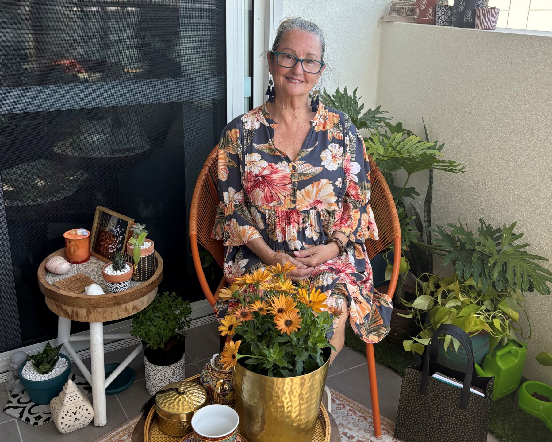 A picture of a woman in a floral print dress wearing glasses sitting in a chair on her balcony surrounded by plants in pots with a cup of tea on a small table beside her.